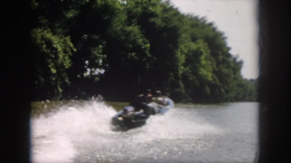 1958 Virginia home-movie frame with the powerboat receding on the river, leaving white spray against a heavily treed shoreline.