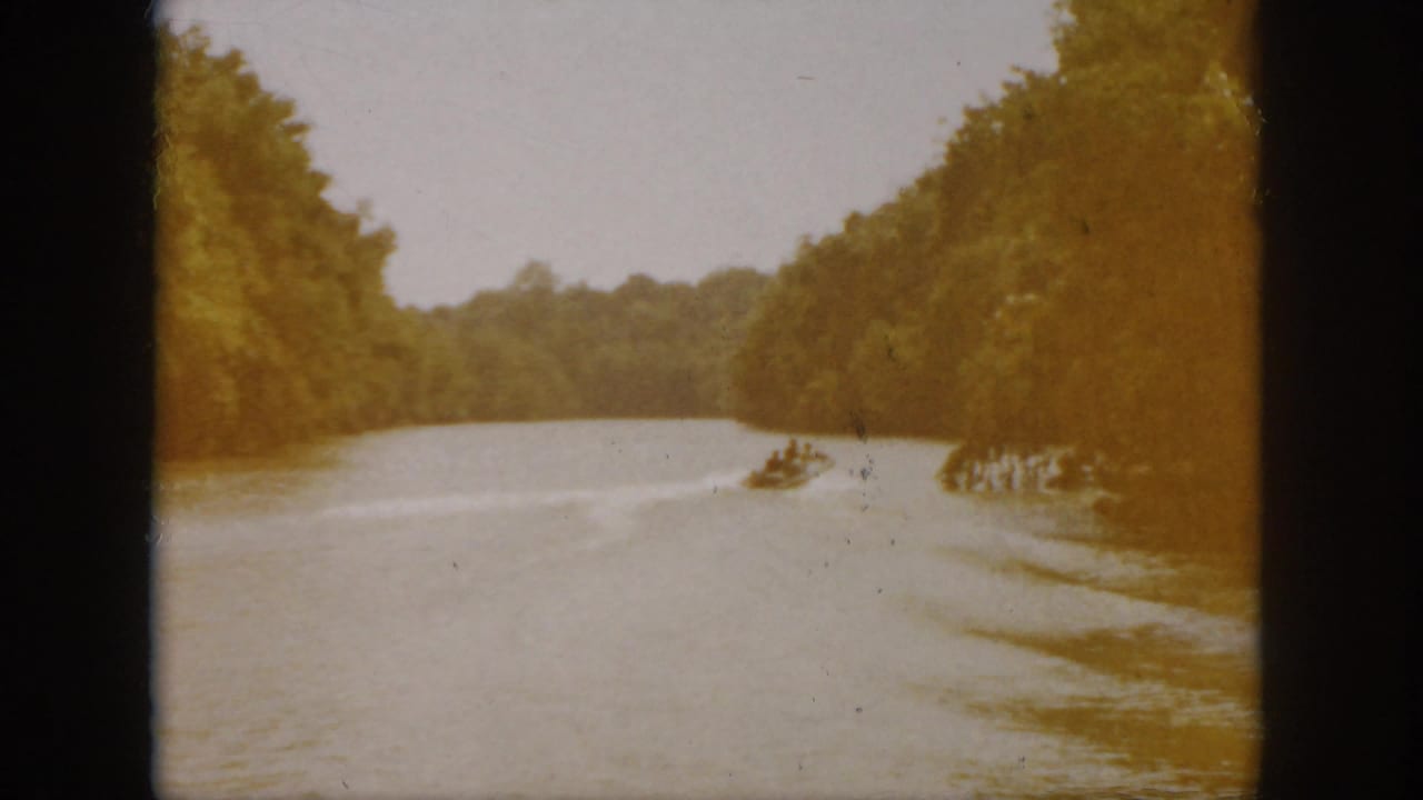 1958 Virginia home-movie frame with a distant powerboat leaving a wake across a wooded river landscape.