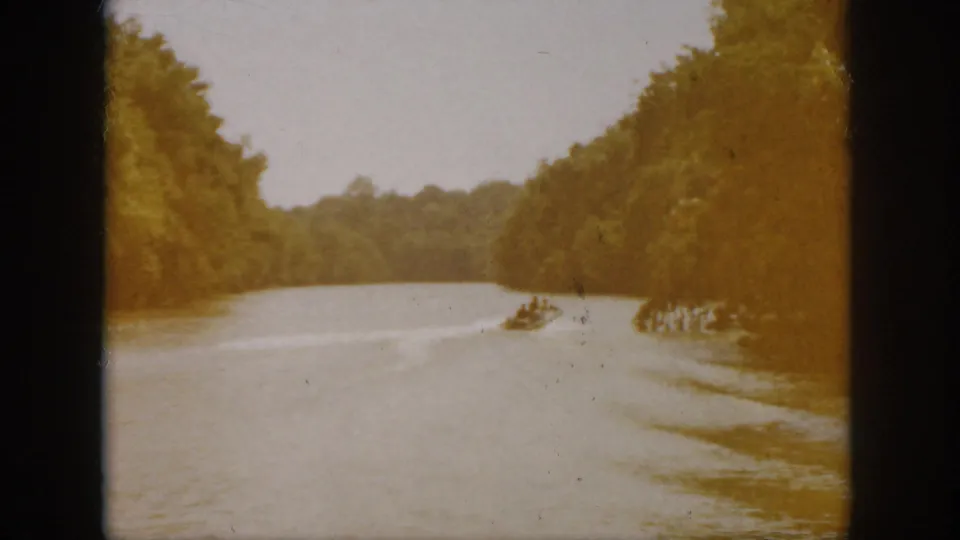 1958 Virginia home-movie frame with a distant powerboat leaving a wake across a wooded river landscape.