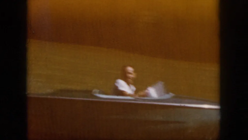 1958 Virginia river scene with a person seated at the helm of a small motorboat, framed softly on faded 8mm film.