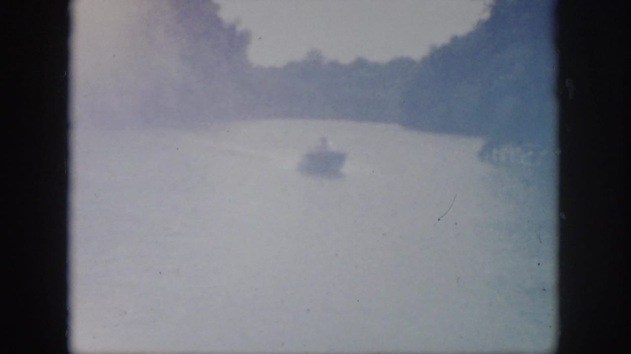 1958 Virginia home-movie frame showing a small motorboat far down a tree-lined river in hazy, overexposed light.