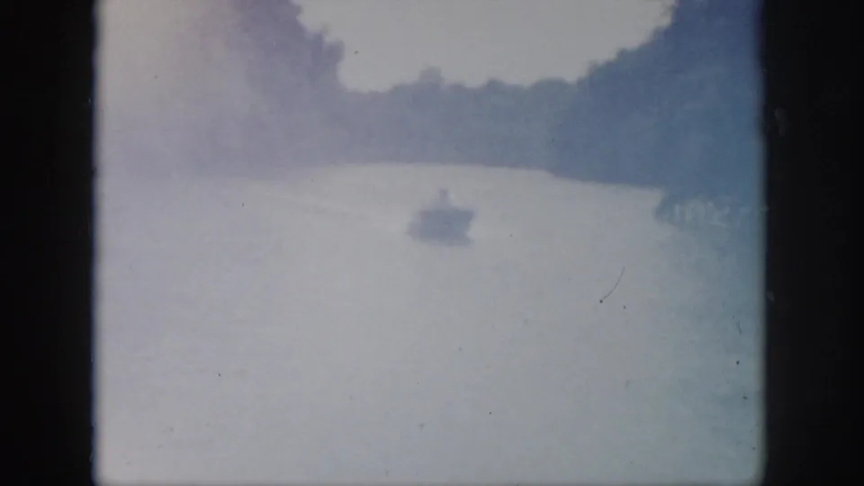 1958 Virginia home-movie frame showing a small motorboat far down a tree-lined river in hazy, overexposed light.
