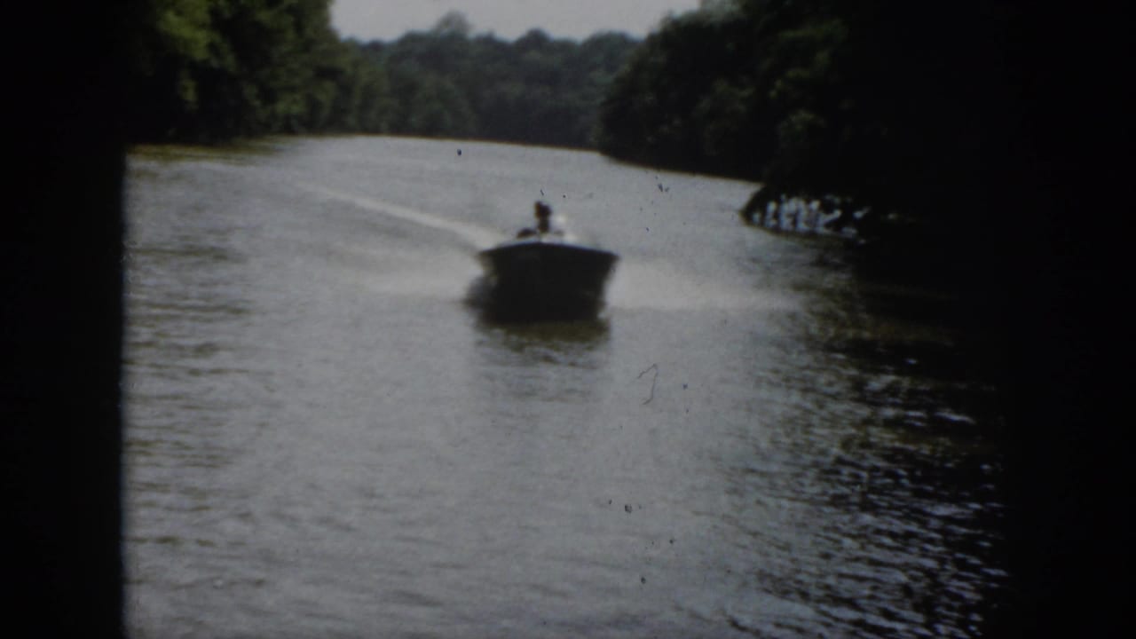 Small motorboat approaches on a wooded river, leaving a white wake behind it.