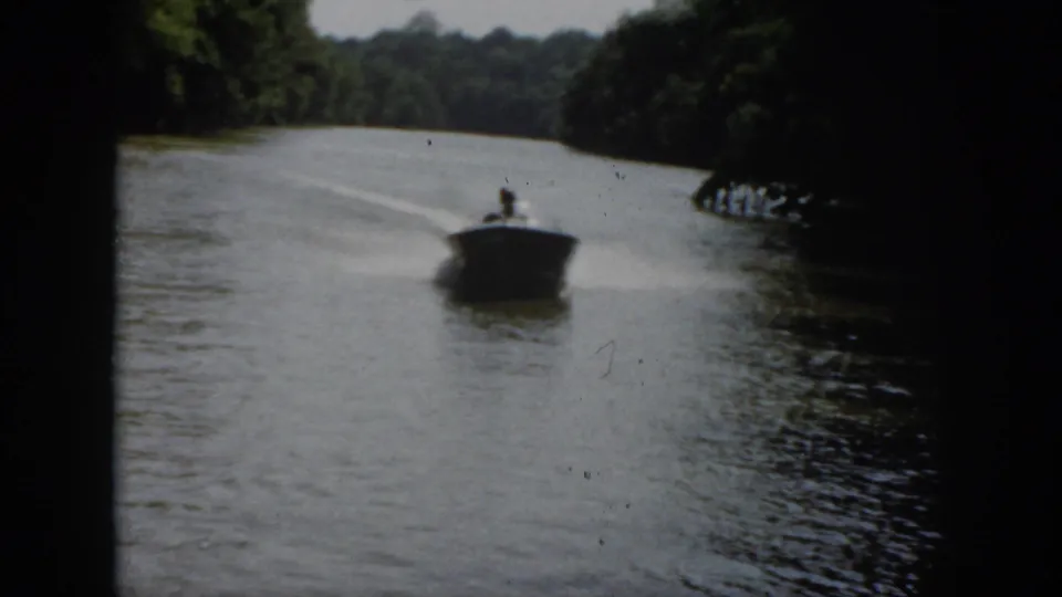 1958 Virginia frame of a small motorboat speeding toward the camera on a wooded river with wake trailing behind.