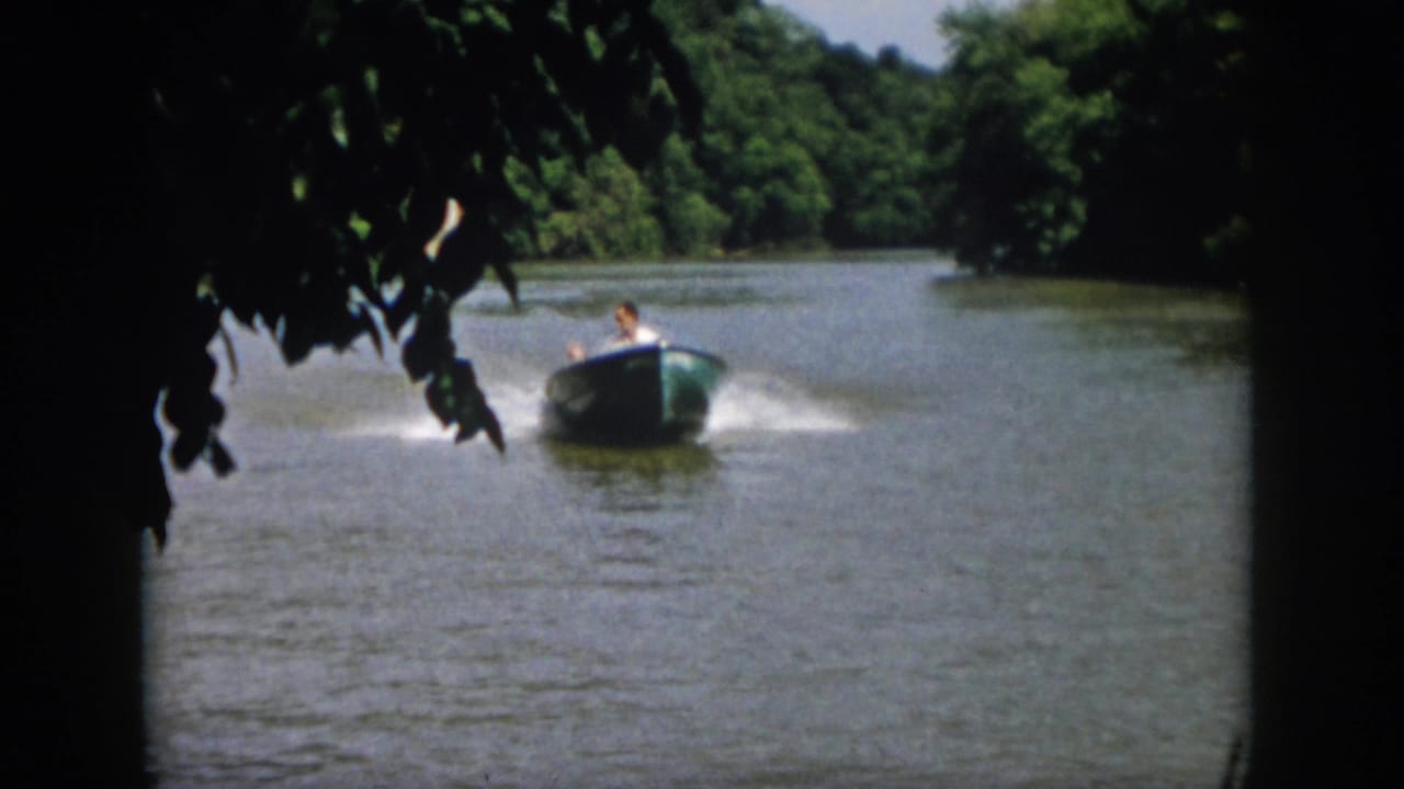 1958 Virginia river frame showing the small motorboat moving away or across the water under leafy branches, with wooded banks behind.