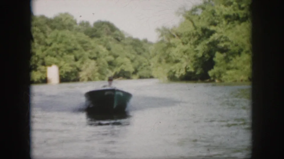 1958 Virginia footage frame with a small motorboat moving away or across a wooded river, its driver faintly visible above the bow.