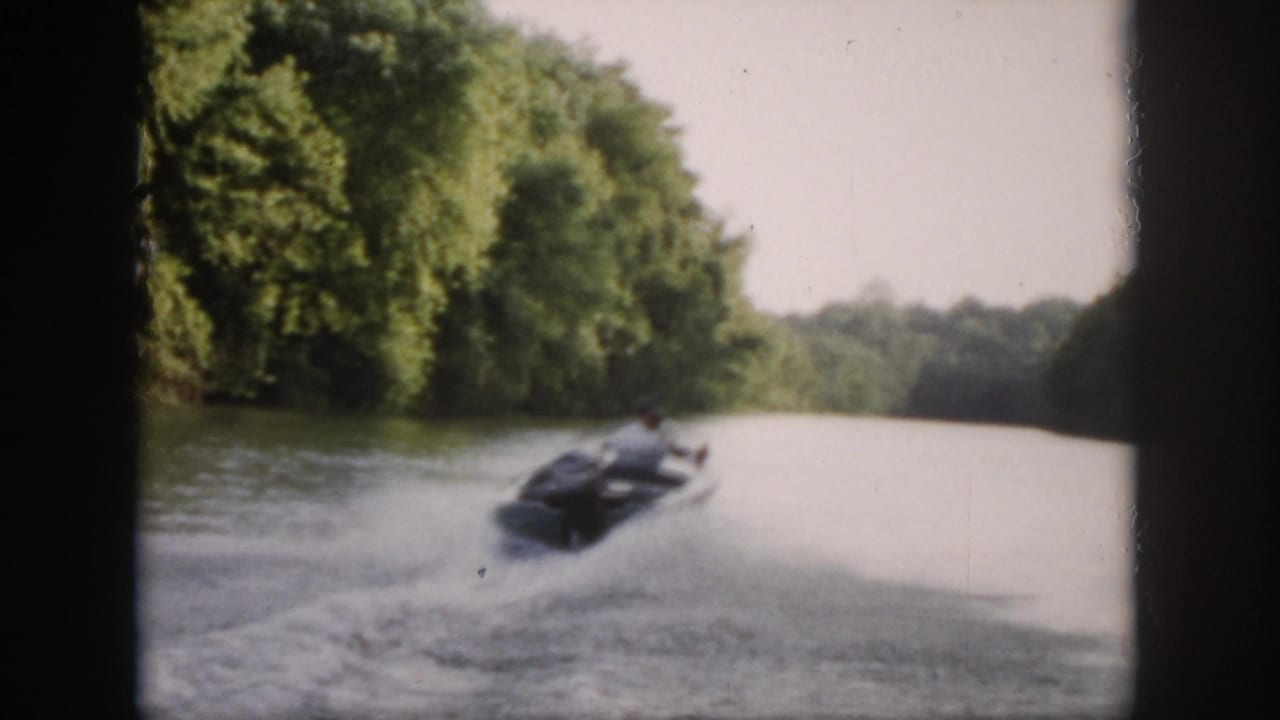1958 Virginia frame showing a small motorboat moving away down a wooded river, with a broad white wake spreading across the water.