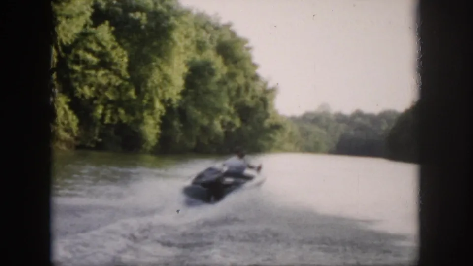 1958 Virginia frame showing a small motorboat moving away down a wooded river, with a broad white wake spreading across the water.