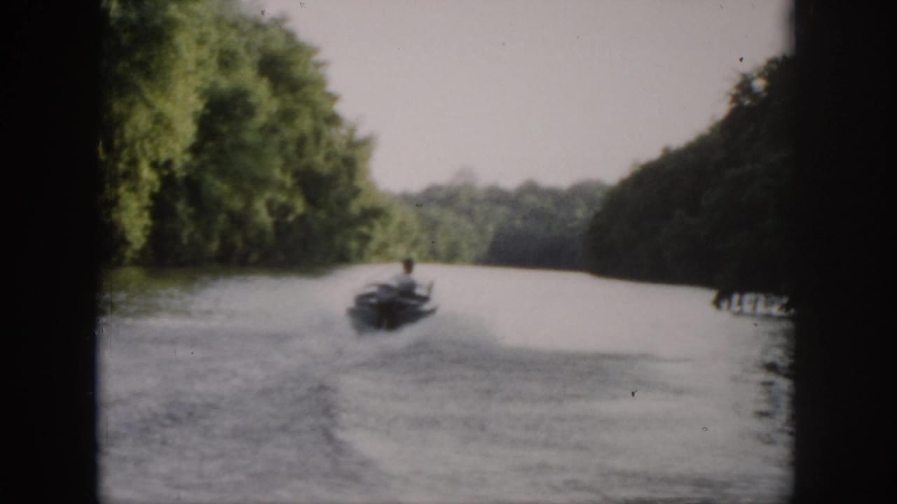 In 1958 Virginia home-movie footage, a small motorboat heads away down a wooded river, leaving a pale wake in soft focus.