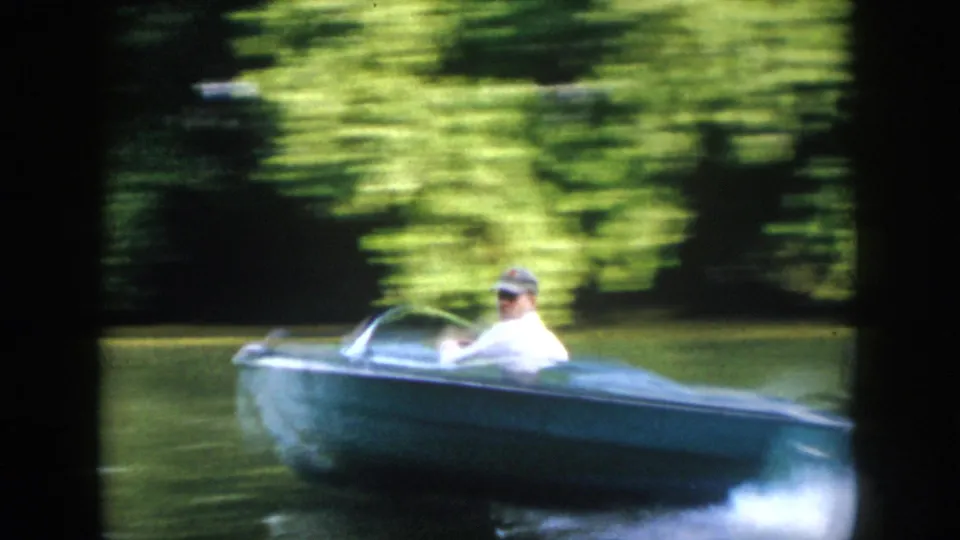 A capped adult driver passes close by in a small blue motorboat, creating spray on the Virginia river in 1958 8mm footage.