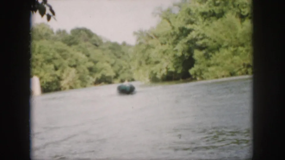 1958 Virginia 8mm frame showing a small green motorboat moving across a calm wooded river at a distance.