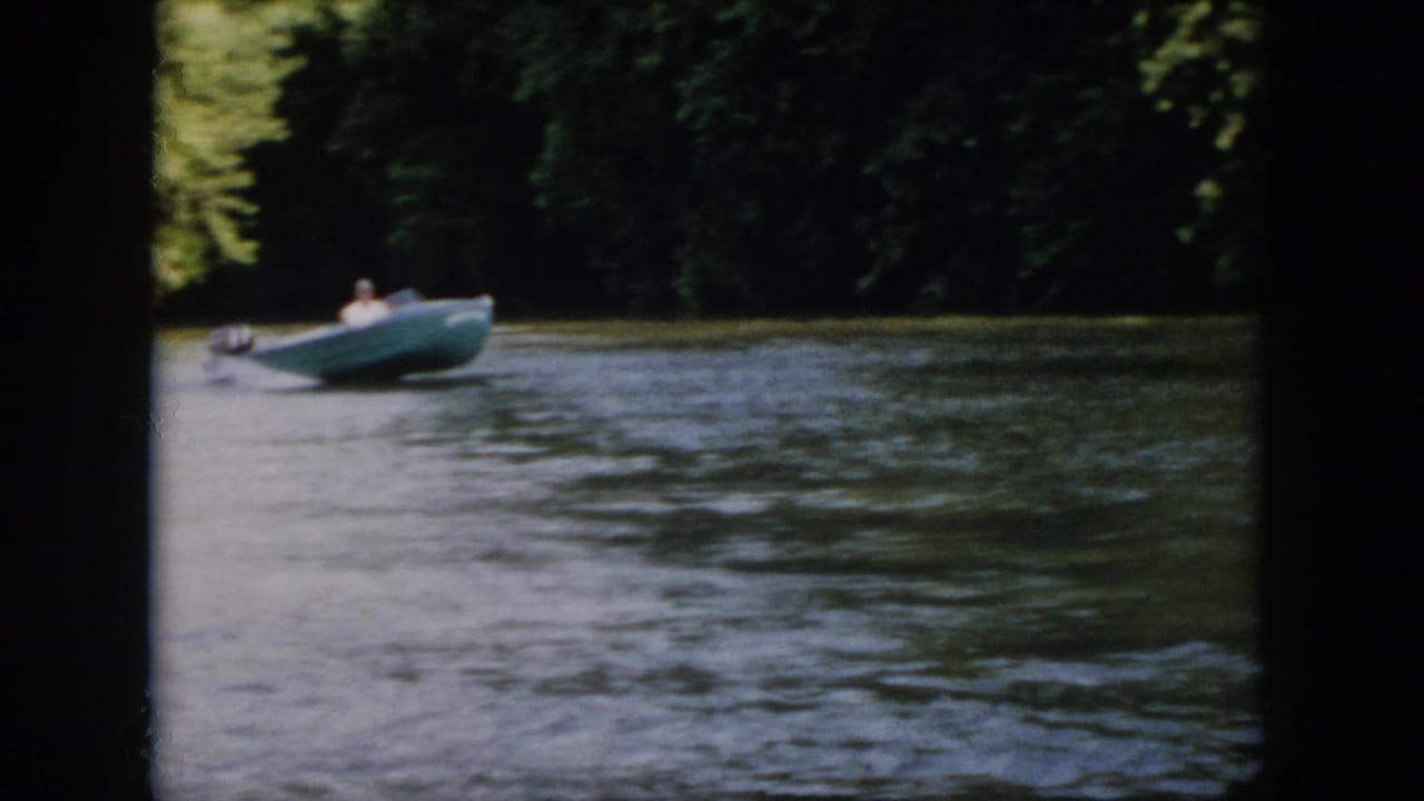 1958 Virginia home-movie still of a man seated in a green motorboat as it moves across rippled river water.