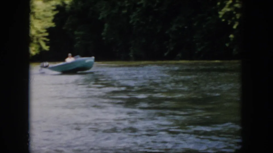1958 Virginia home-movie still of a man seated in a green motorboat as it moves across rippled river water.