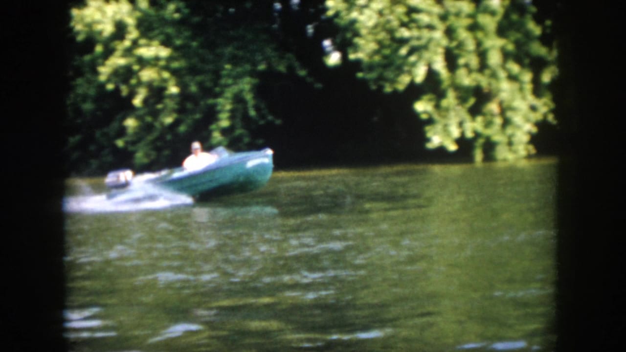 A man steers a green motorboat on a wooded Virginia river, leaving a bright white wake in 1958 8mm footage.