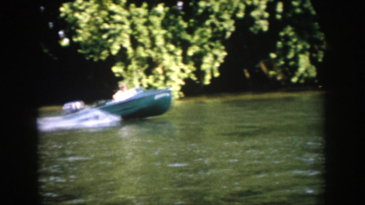 1958 Virginia 8mm frame showing a green motorboat crossing a calm wooded river with a man seated at the controls.