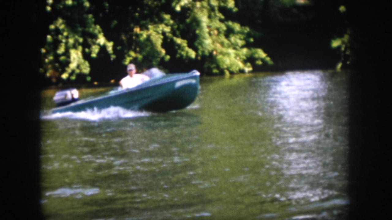 Green motorboat speeds along the river near overhanging trees, throwing a low wake.
