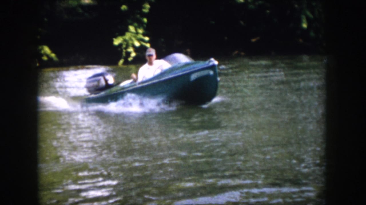 A man in a light shirt and cap steers the green outboard motorboat across the wooded Virginia river.