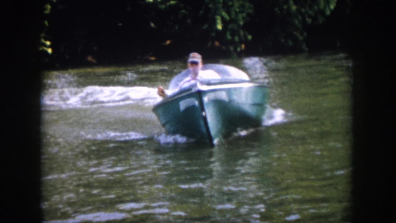 The green motorboat turns toward camera on the wooded river, its bow and windshield visible as wake spreads behind it.