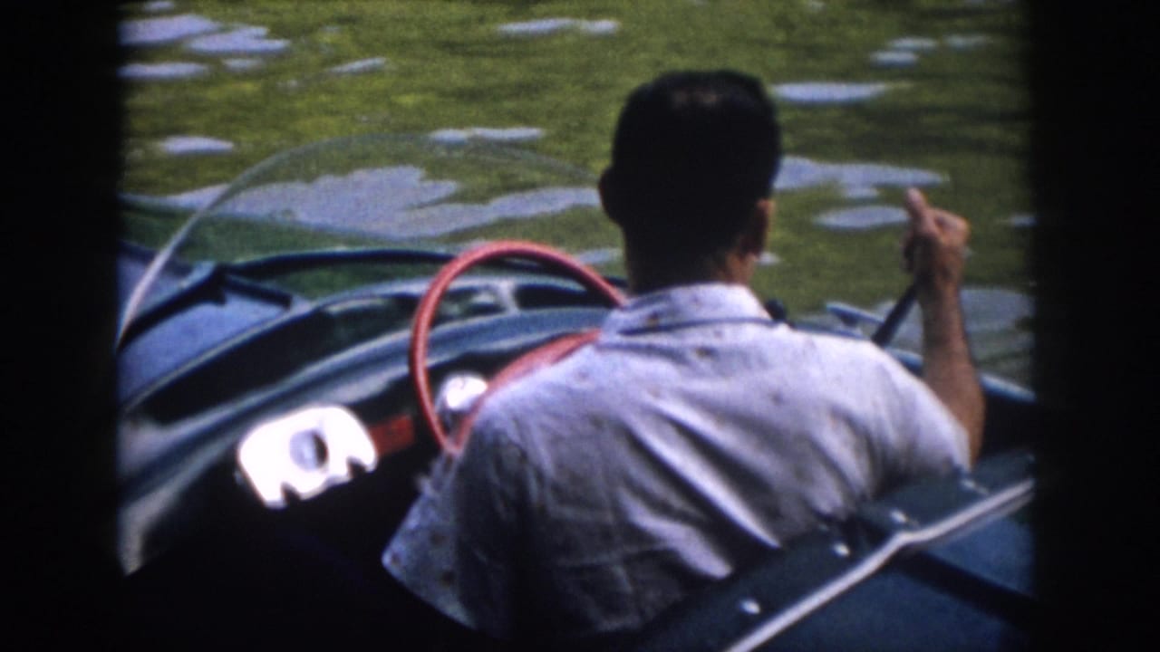 1958 Virginia home-movie frame of a man in a light shirt steering a green speedboat across rippled lake water.