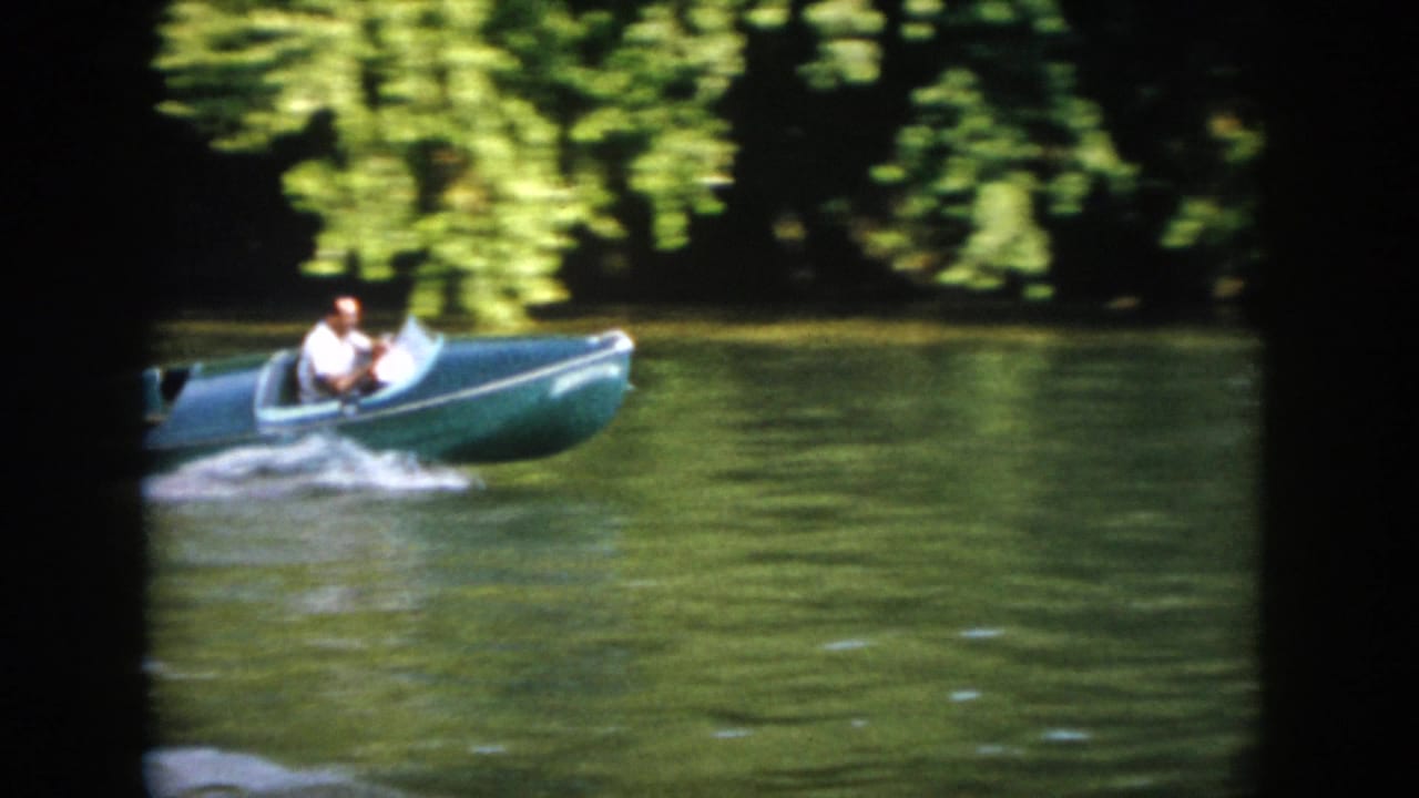 1958 Virginia frame of the green speedboat passing across sunlit lake water with trees along the far shore.