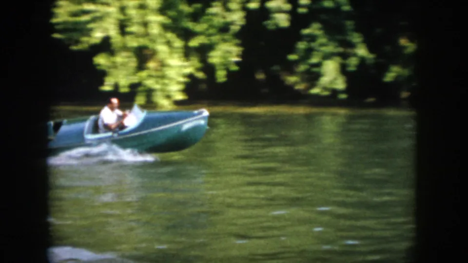 1958 Virginia frame of the green speedboat passing across sunlit lake water with trees along the far shore.