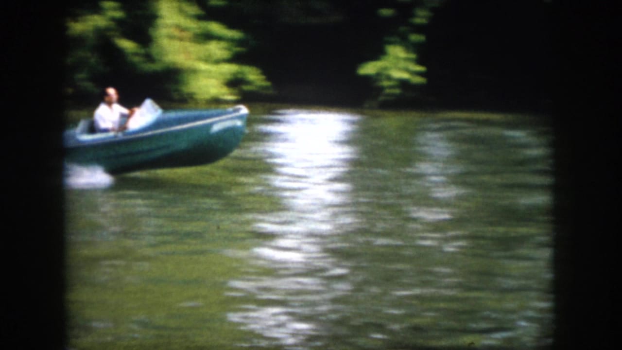 1958 Virginia home-movie frame of a man in a small blue motorboat turning on a calm, wooded river.