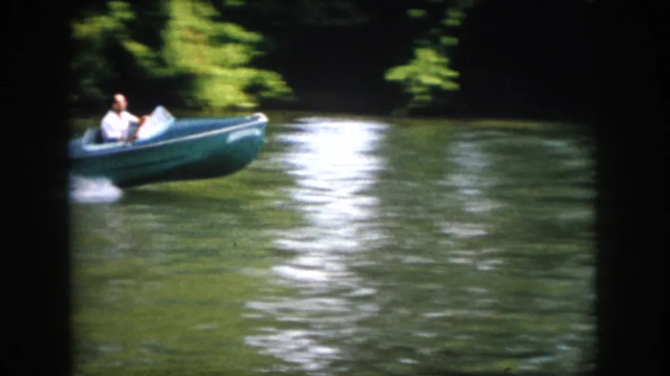 1958 Virginia home-movie frame of a man in a small blue motorboat turning on a calm, wooded river.