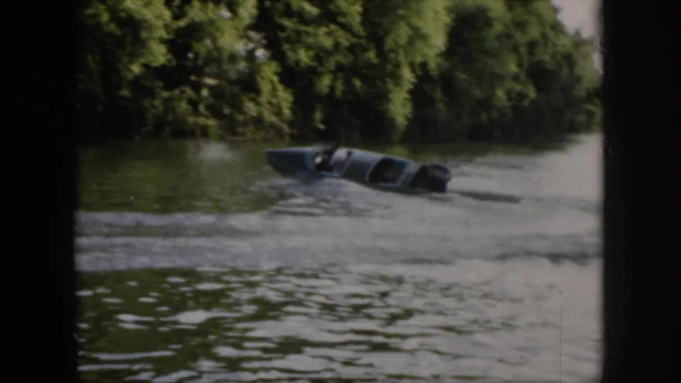 1958 Virginia frame of a small blue motorboat leaning into a circular turn on a wooded river.
