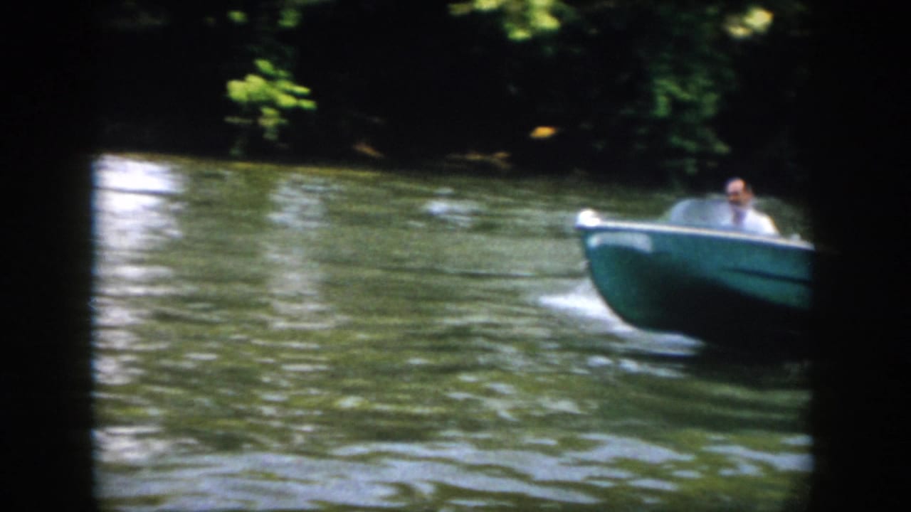 1958 Virginia frame with a blue motorboat and adult driver passing across calm green river water.