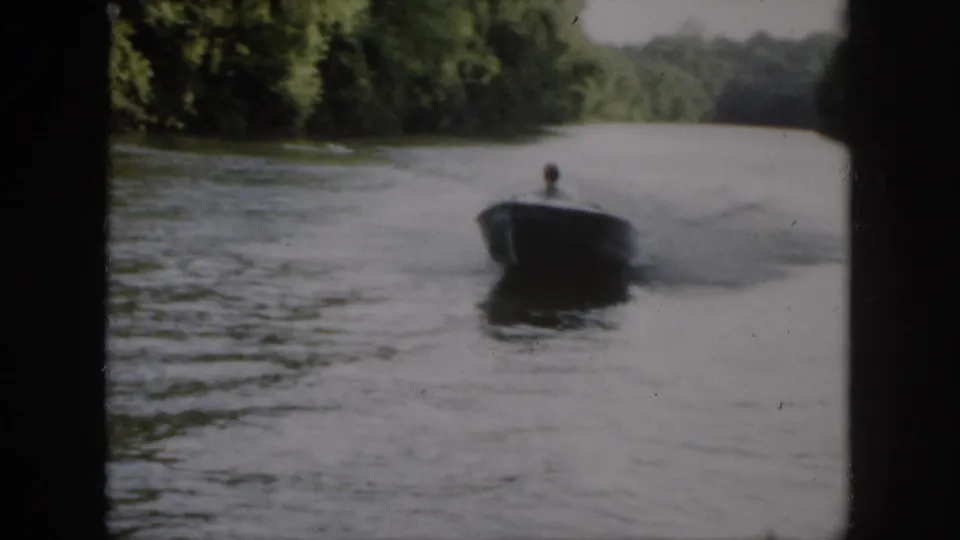 In 1958 Virginia home-movie footage, a small motorboat approaches along a tree-lined waterway with one adult visible aboard.