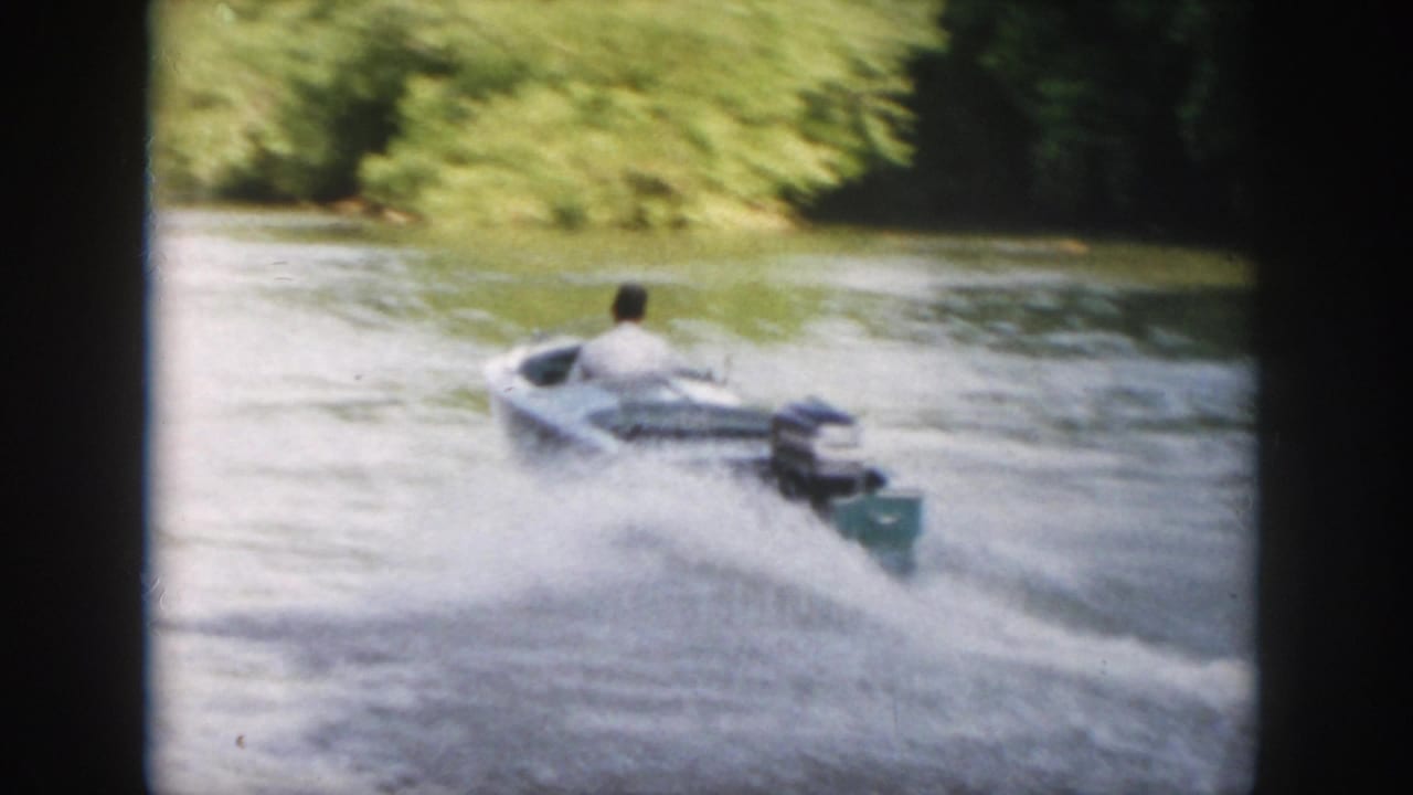 A man in a light shirt pilots a small motorboat away from the camera, leaving spray and wake on the Virginia waterway.