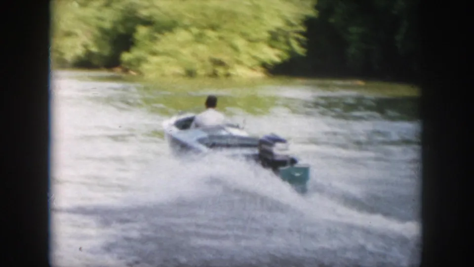 A man in a light shirt pilots a small motorboat away from the camera, leaving spray and wake on the Virginia waterway.