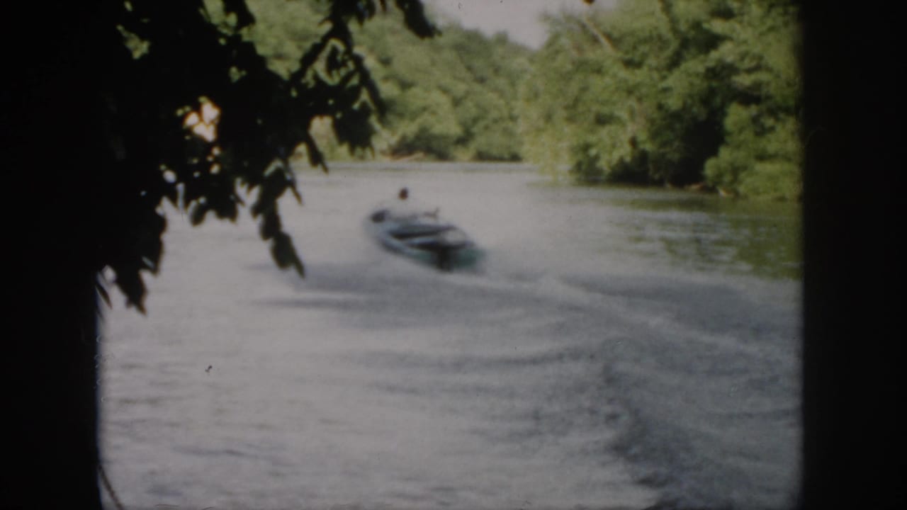 The motorboat recedes along a tree-lined Virginia river, with overhanging leaves framing the home-movie shot.