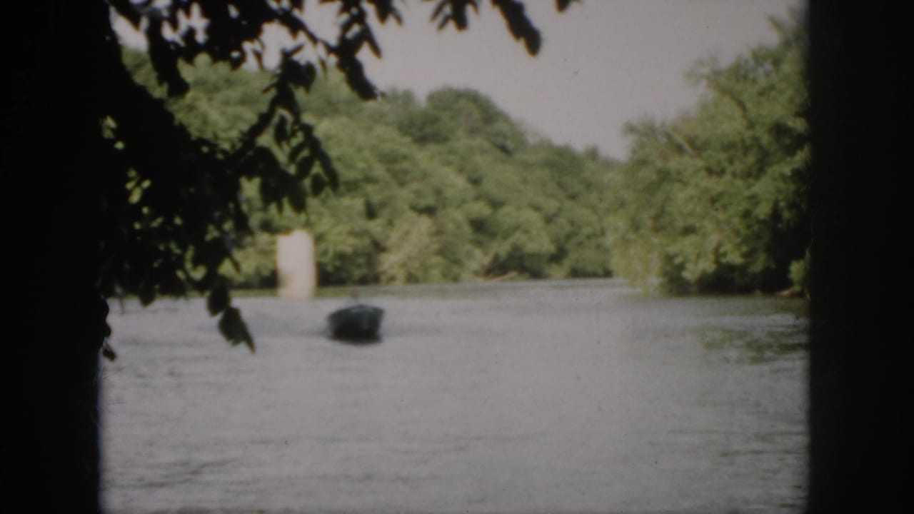 1958 Virginia 8mm frame showing a small motorboat at a distance on a tree-lined river, partly framed by dark overhanging branches.