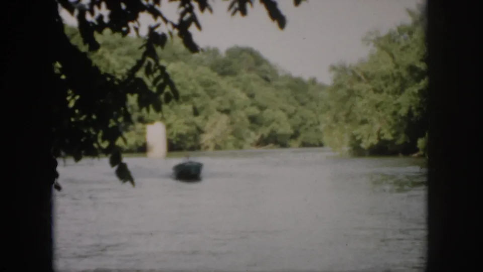 1958 Virginia 8mm frame showing a small motorboat at a distance on a tree-lined river, partly framed by dark overhanging branches.