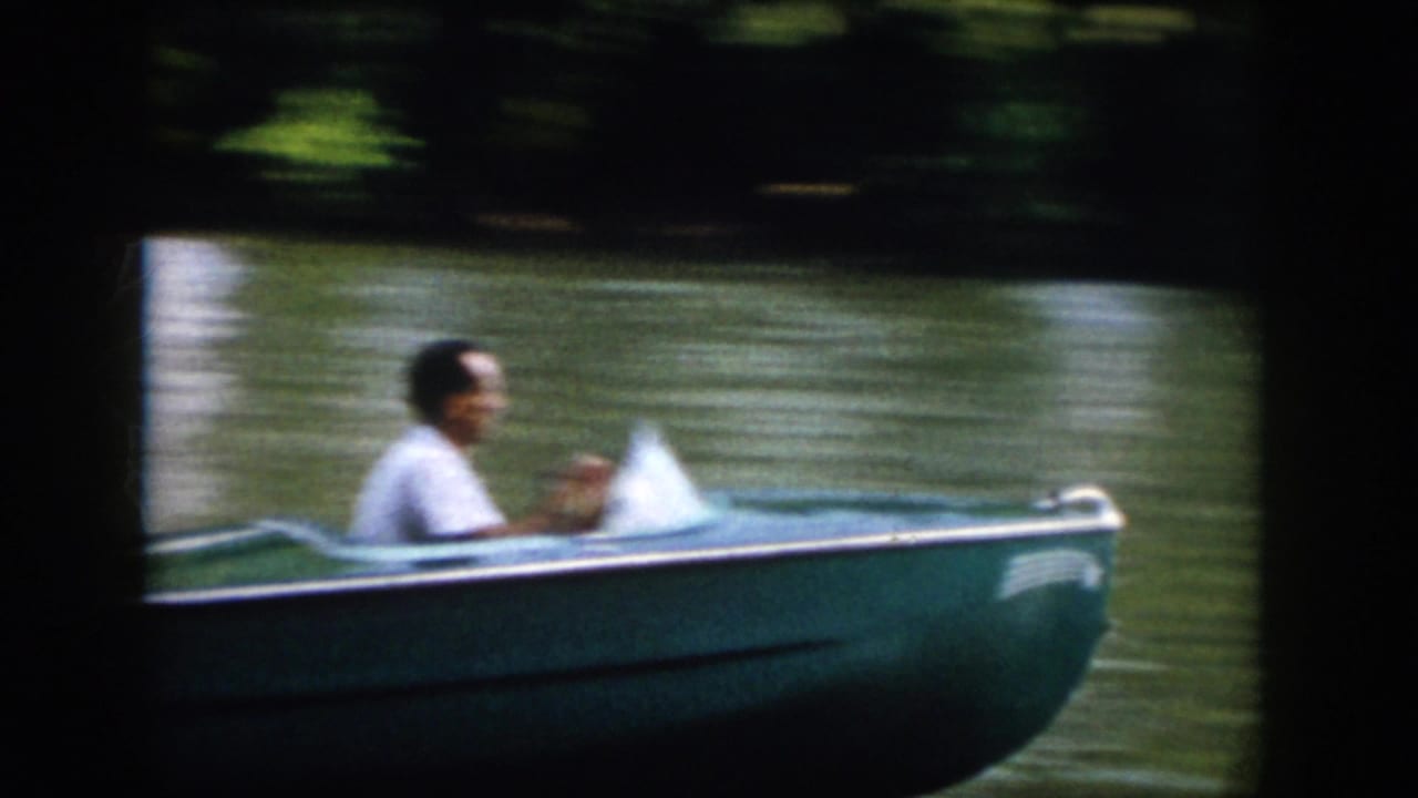Adult man in a light shirt pilots a small blue boat past the camera on a Virginia river in 1958 8mm footage.