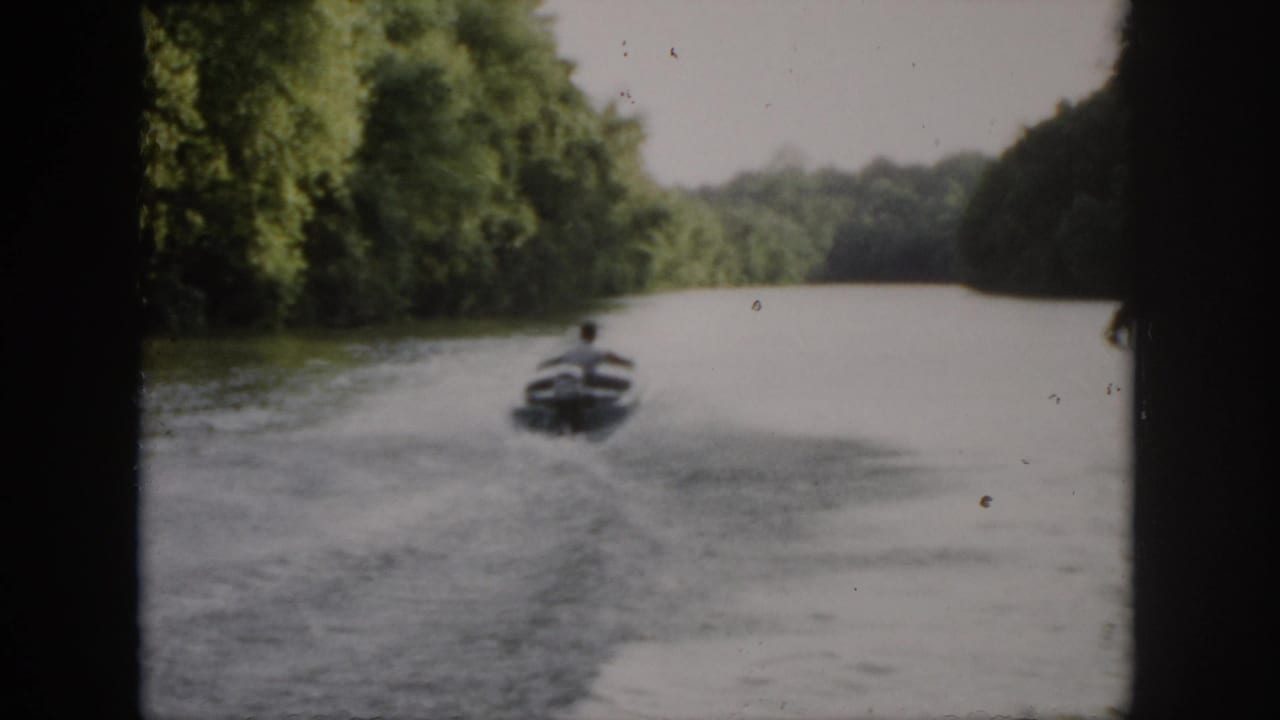 1958 Virginia home-movie frame showing a small speedboat receding across green water between wooded banks.