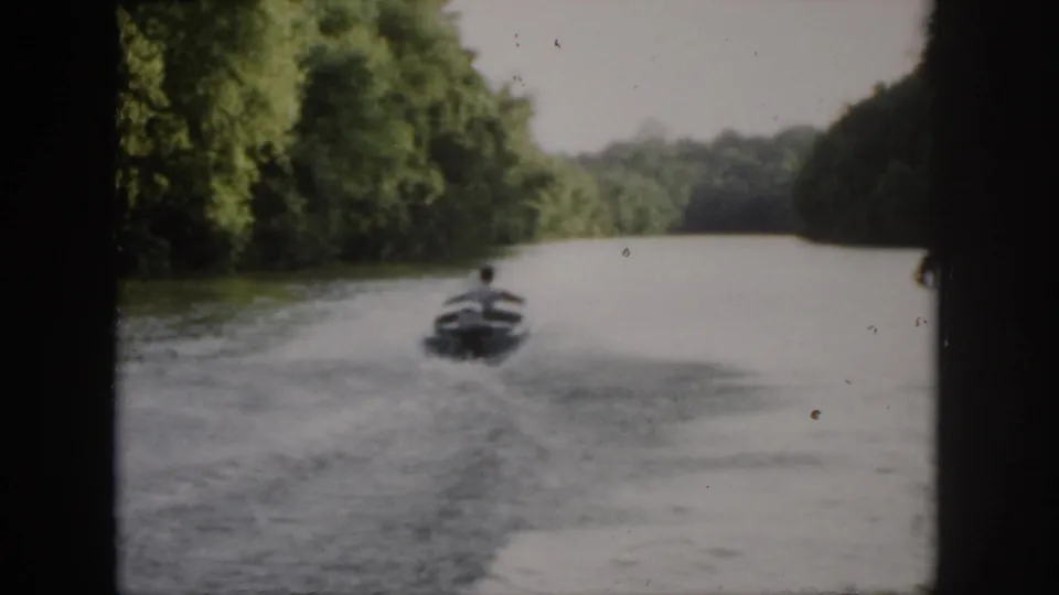 1958 Virginia home-movie frame showing a small speedboat receding across green water between wooded banks.