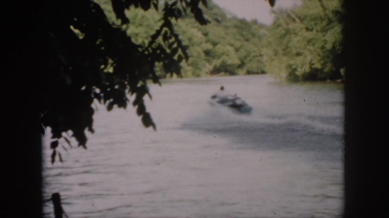 1958 Virginia 8mm frame of a small motorboat moving across a wooded river, seen through dark foreground branches.