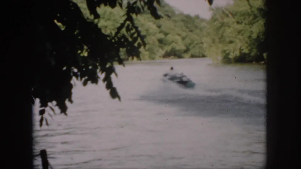 1958 Virginia 8mm frame of a small motorboat moving across a wooded river, seen through dark foreground branches.
