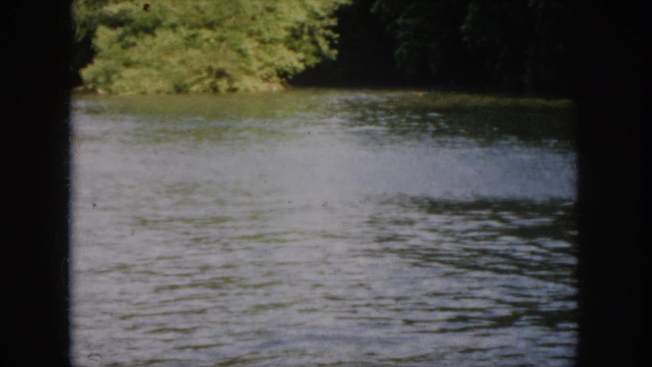 1958 Virginia home-movie frame showing calm river water, wooded bank, and visible 8mm frame edges before the boat enters.