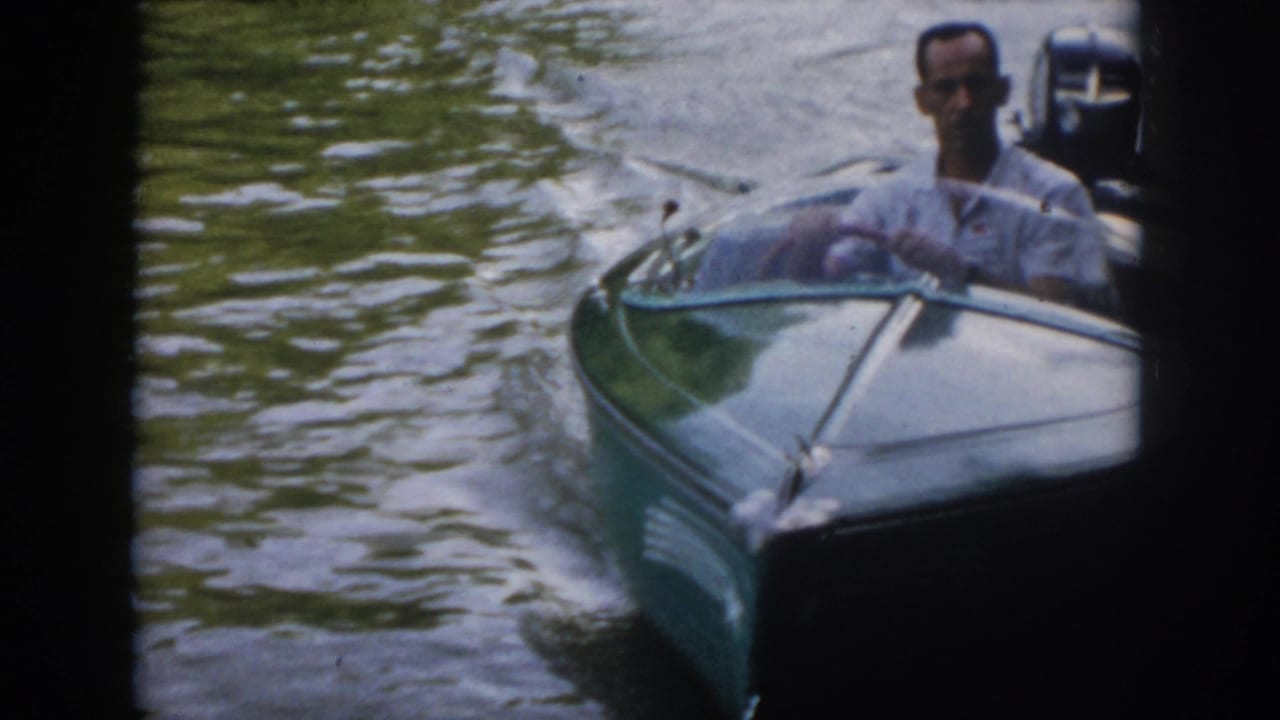 Close 1958 Virginia frame of a man piloting a small speedboat past the camera, with outboard motor, wake, and river water visible.
