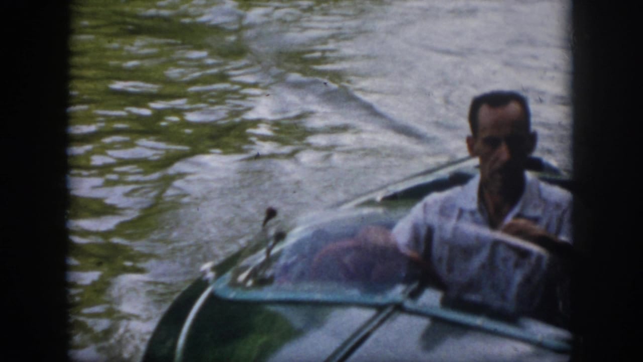 1958 Virginia home-movie frame showing a man in a light shirt seated at a small motorboat as it moves through river water.