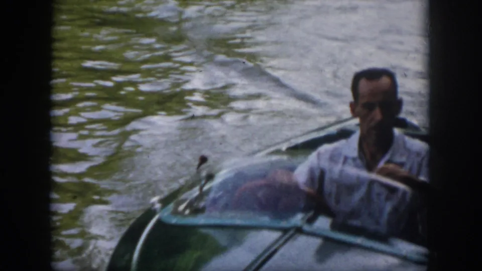 1958 Virginia home-movie frame showing a man in a light shirt seated at a small motorboat as it moves through river water.