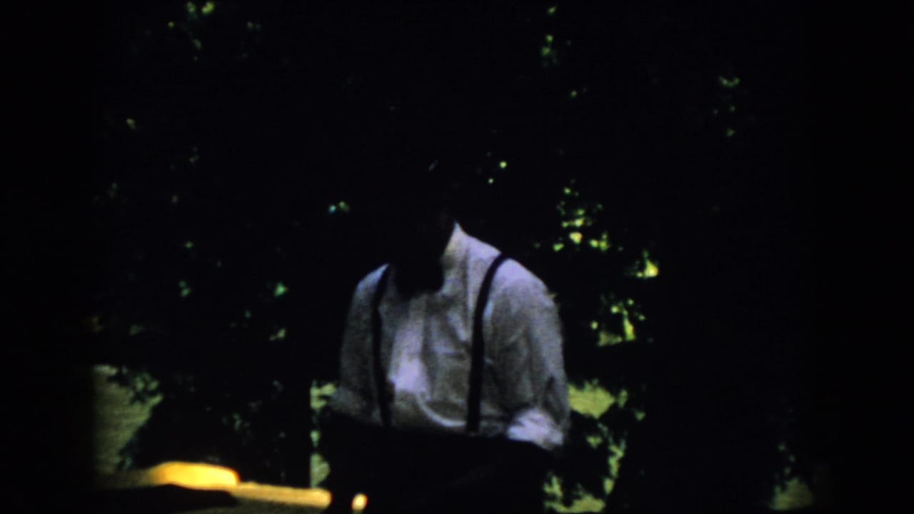 Dark 1958 Virginia home-movie frame showing a man in a light shirt and suspenders standing outdoors under trees.