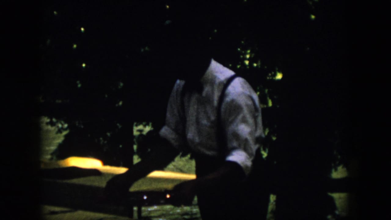 1958 Virginia home-movie frame of a man in suspenders near a yellow-lit surface, with dark foliage and faint lights behind him.