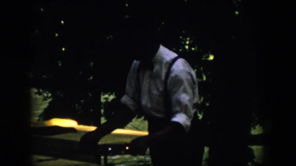 1958 Virginia home-movie frame of a man in suspenders near a yellow-lit surface, with dark foliage and faint lights behind him.