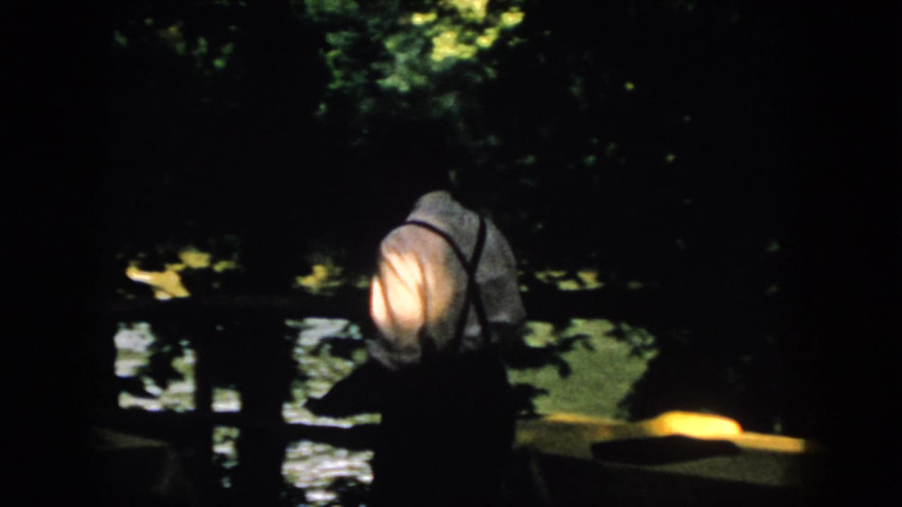Man in suspenders walks near a dark waterfront with trees behind him.