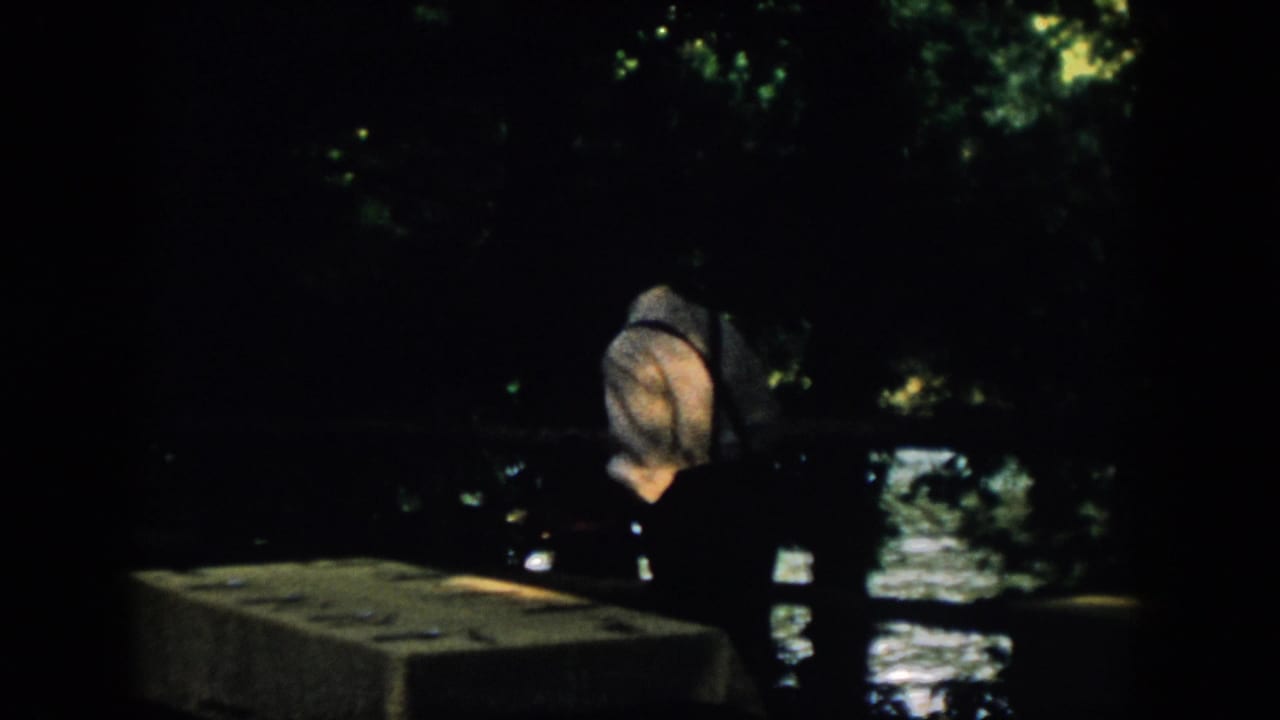 A dark 8mm frame shows the man near a low ledge or wall, with water reflections and tree cover in the background.