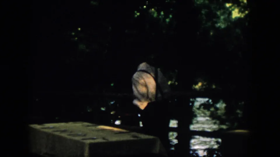 A dark 8mm frame shows the man near a low ledge or wall, with water reflections and tree cover in the background.